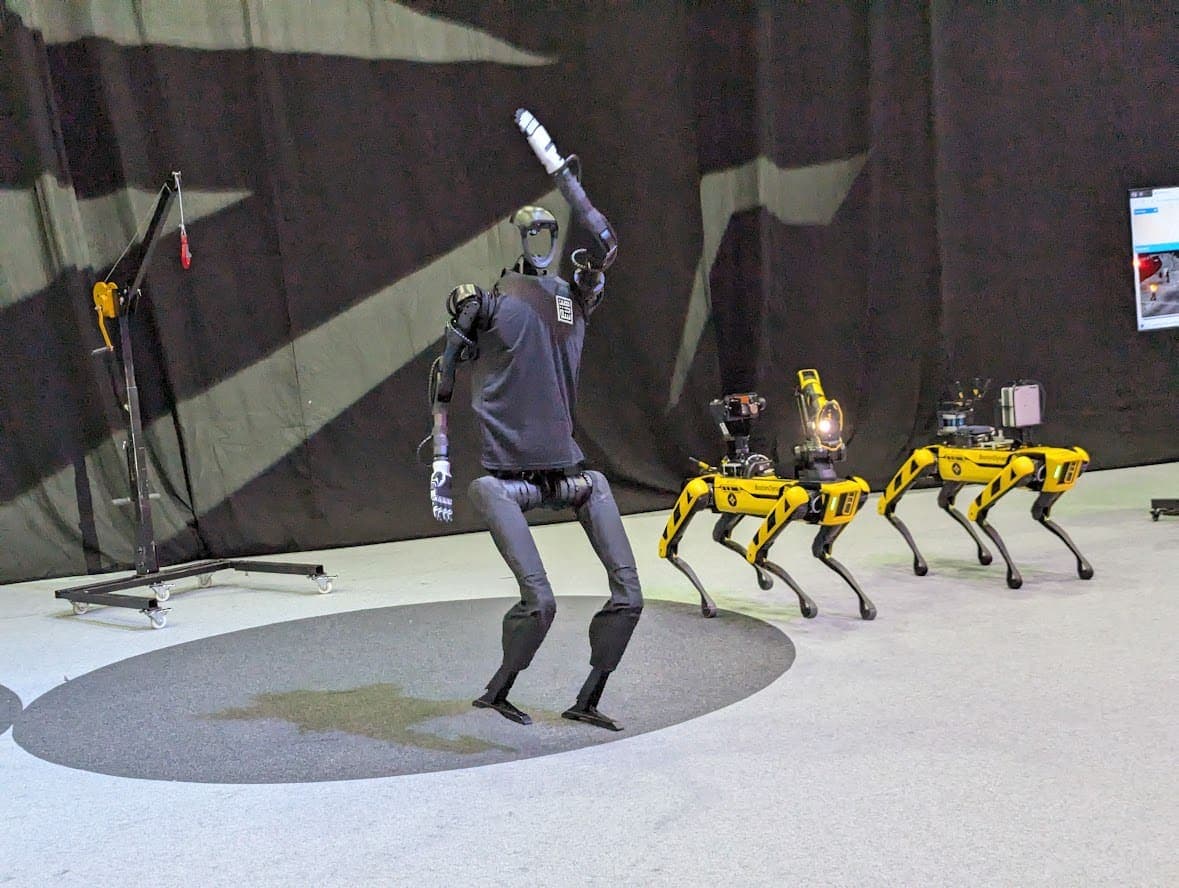 Unitree humanoid robot mid-motion on an exhibition stage, surrounded by a fleet of quadruped robots under stage lighting