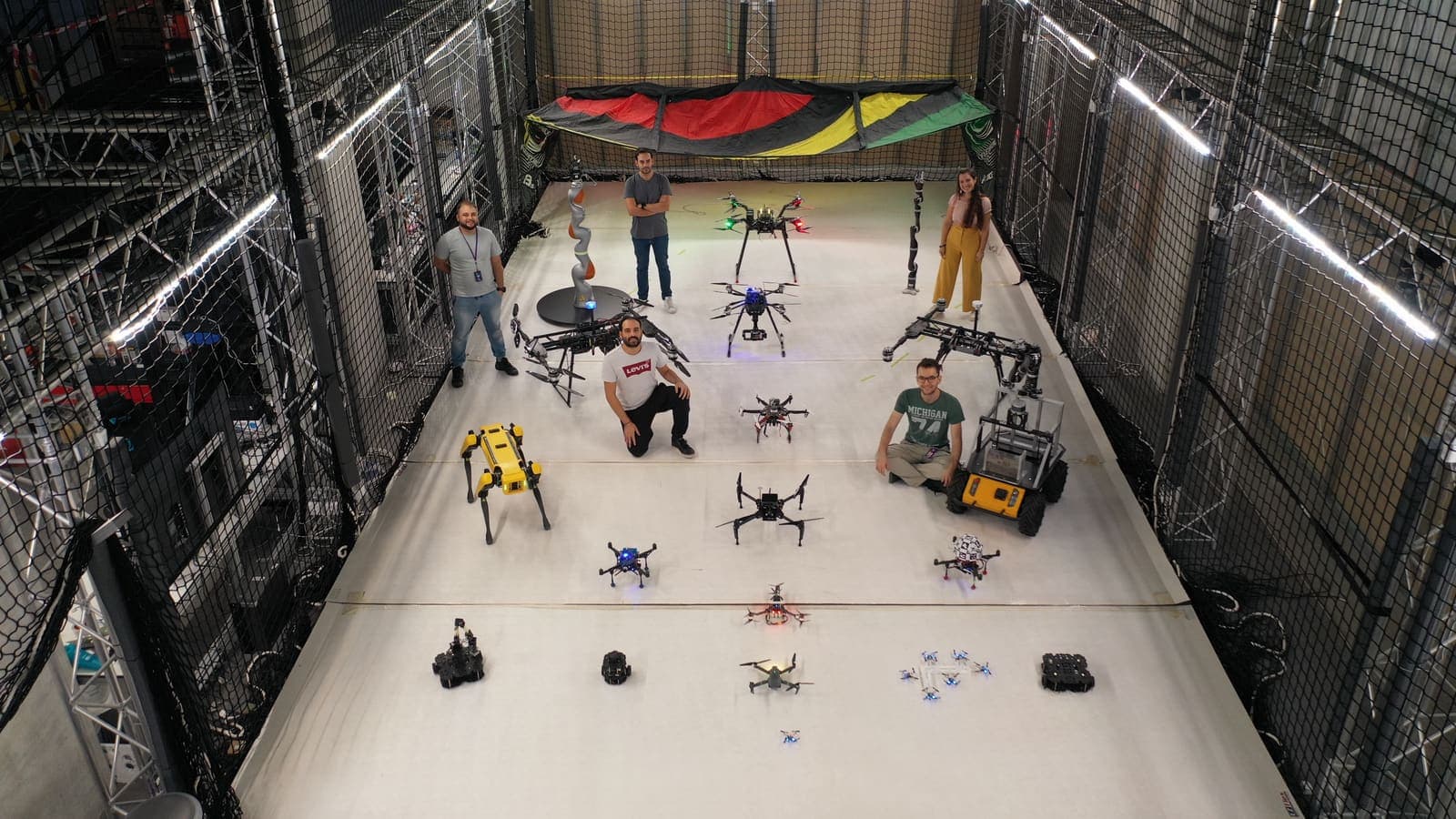 Aerial view of the Kinesis arena floor with a Spot quadruped, a heavy-lift hexacopter, and ground robots staged between truss columns