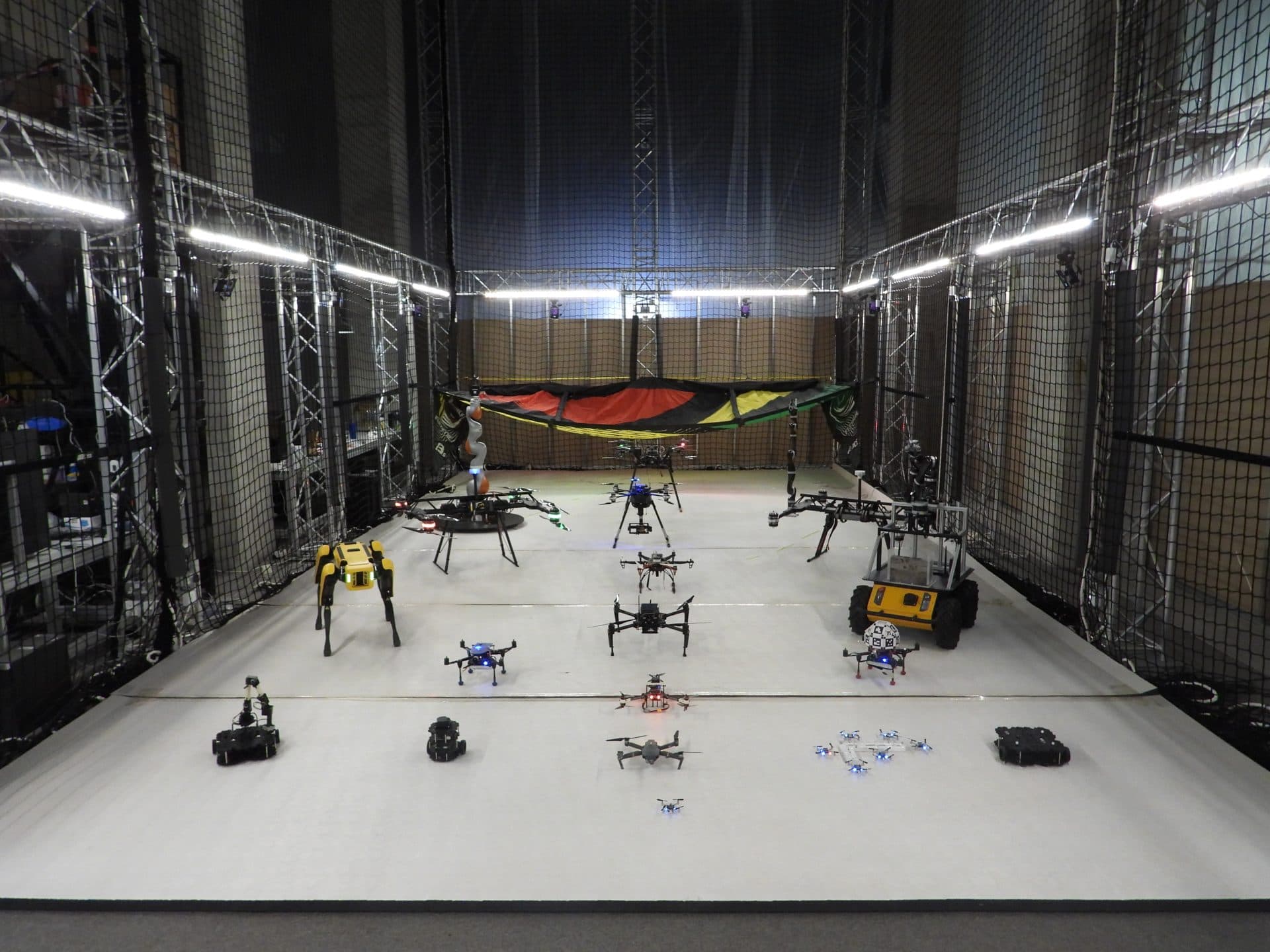 The Kinesis arena enclosed by truss structures and safety nets, with a fleet of drones, a Spot quadruped, ground robots, and a KUKA arm arranged on the floor