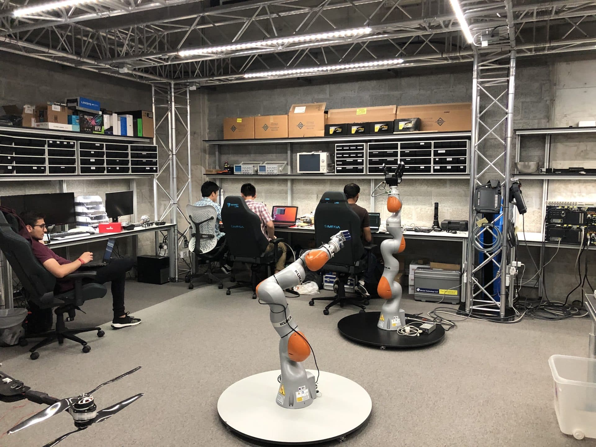The Kinesis workspace with researchers at workstations beneath equipment shelving, and two KUKA collaborative arms on turntables in the foreground