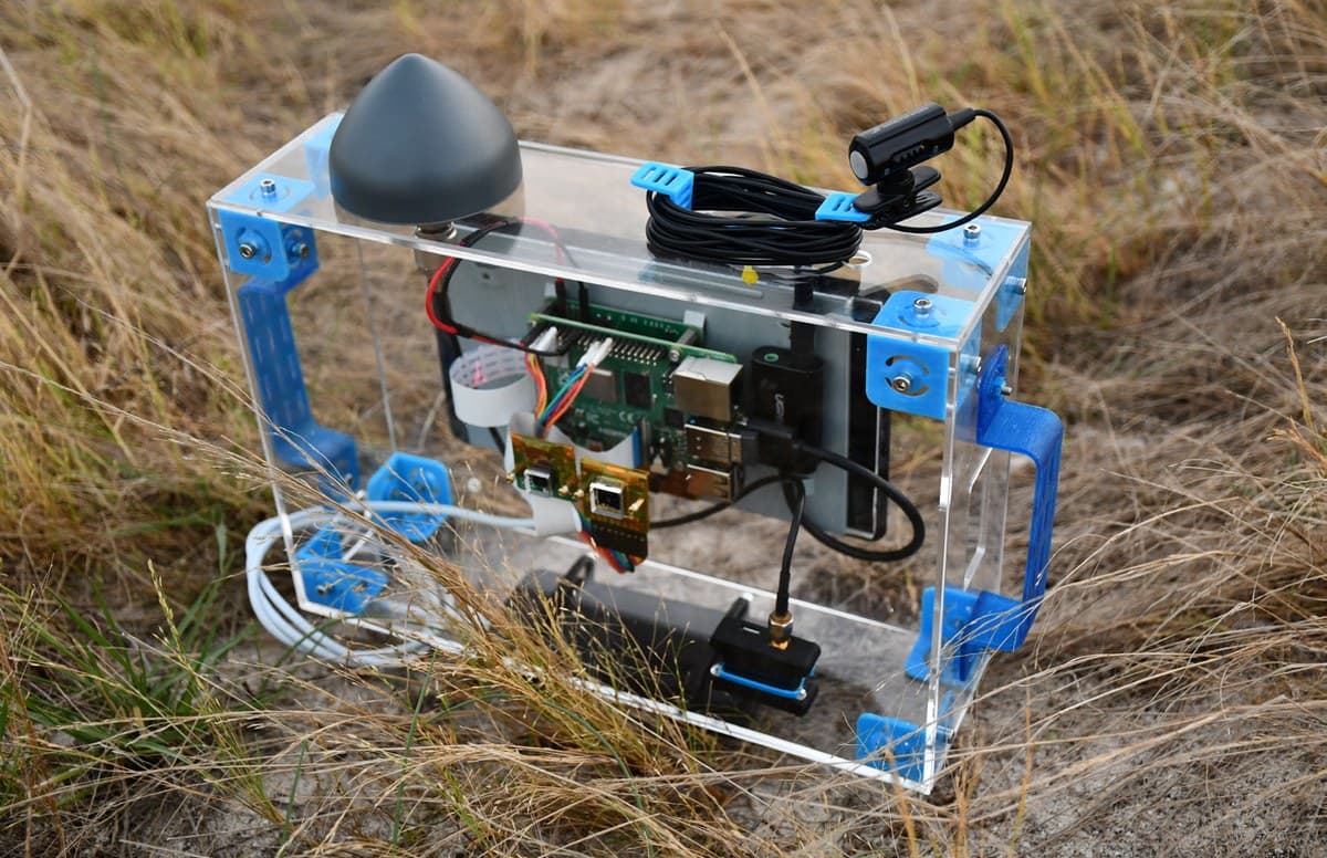 Handheld acrylic sensor-fusion data collector with blue 3D-printed brackets, Raspberry Pi, GPS dome antenna, and clipped contact microphone resting in dry field grass