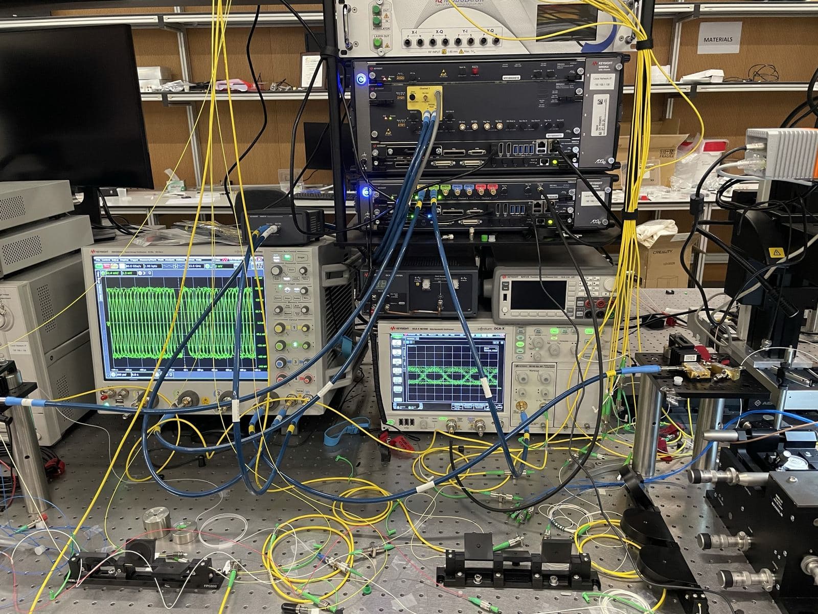 High-speed photonics test stack with Keysight arbitrary waveform generators, Infiniium oscilloscopes showing eye diagrams, and fiber and RF cabling on an optical breadboard