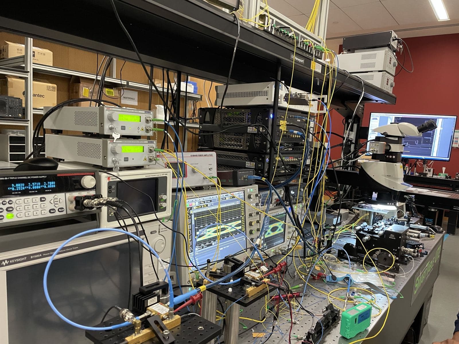 Photonics characterization bench with Thorlabs amplifiers, Keysight instruments, Leica microscope, and dense fiber cabling on a Newport optical table