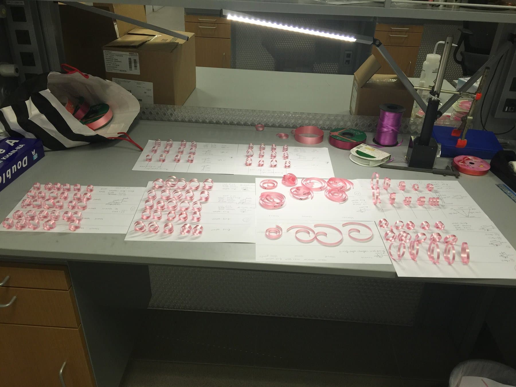 Laboratory workbench covered with pink ribbon curl samples arranged by test condition under a task light